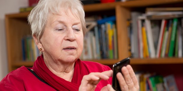 Ein alte Frau mit kurzen grauen Haaren und roten Pullover sitzt vor einem Bücherregal. Sie hat ein Smartphone in der Hand und schaut darauf.