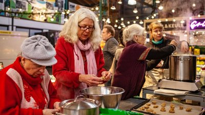 Drei alte Frauen und eine junge Frau stehen in einer Markthalle und kochen dort gemeinsam.