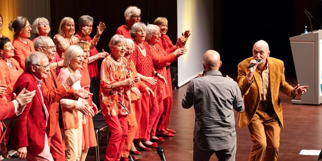 Auf einer Bühne: Ein Chor mit vielen alten Menschen. sie sind in Rottönen gekleidet. Vor ihnen ein Solist mit Mikrofon in der Hand. Er schaut den Dirigenten an.