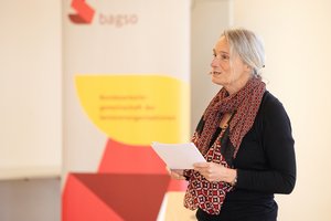 Die Modratorin, Katja Nellissen, eine Frau mittleren Alters, steht auf einer Bühne. Sie hat einen schwarzen Pulli an und hat ein buntes Tuch um den Hals. Sie hält Notizen in den Händen und hat ein Headset an. Im Hintergrund steht ein Banner der BAGSO. Klick öffnet eine vergrößerte Ansicht.