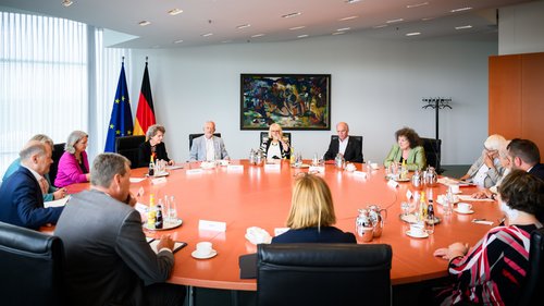 14 Personen sitzen um einen runden Konferenztisch aus Kirschbaumholz. Im Hintergrund sieht man die Deutschland- und Europafahne und ein abstraktes Gemälde. 
