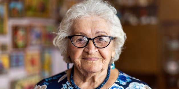 Alte Frau mit grauen Haaren. Sie trägt eine Brille, eine blaue Halskette und Hängeohrringe. Sie hat eine blaue Bluse mit Blumenmuster an.