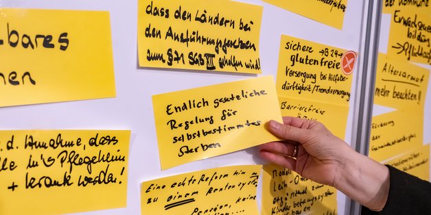 Eine Pinwand, auf der beschriftete gelbe Zettel kleben. Eine Notiz wird gerade von einer Hand angeheftet.