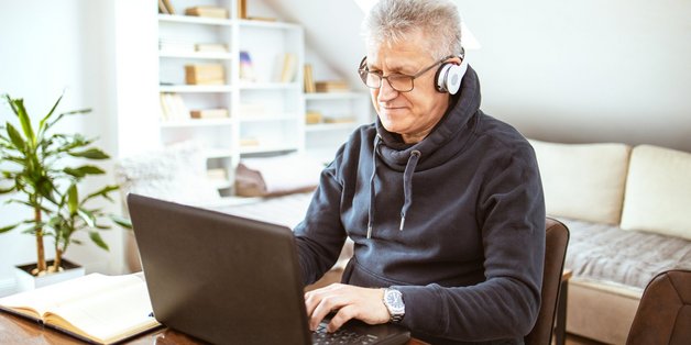 Älterer Mann mit Headset sitzt in seinem Wohzimmer vor einem Laptop