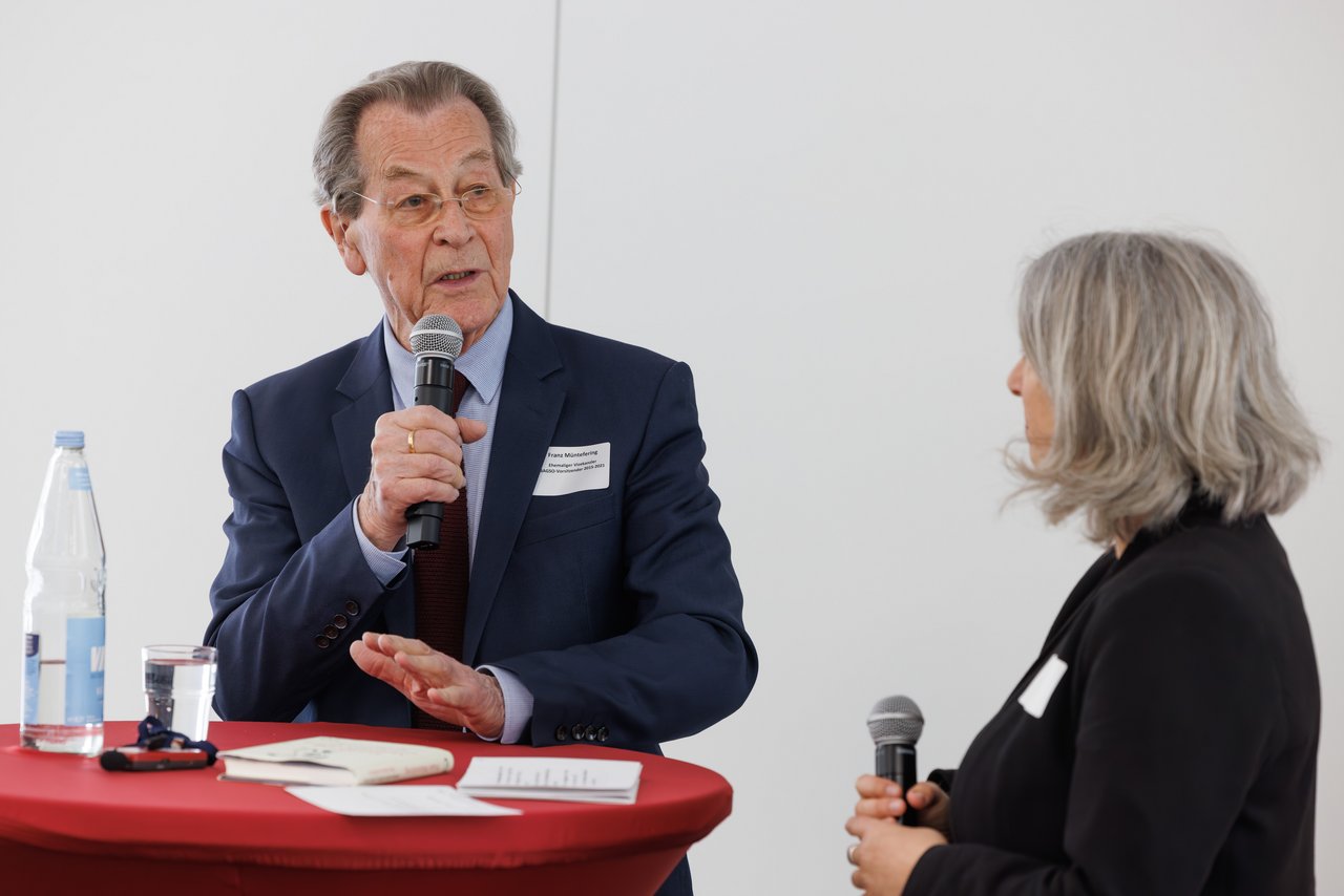 Franz Müntefering, alter Mann mit Mikro in der Hand. Er steht mit einer Frau, mittleren Alters  an einem Stehtisch mit roter Tischdecke. Sie hält auch ein Mikro in der Hand und hört ihm zu.