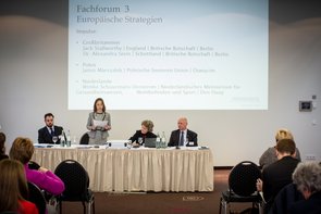 An einem Rednertisch sind zwei Frauen und zwei Männer zu sehen. Eine Frau steht gerade und liest etwas von einem Blatt Papier ab. Auf dem Tisch sind Unterlagen eine Laptop und Getränke zu sehen. Klick öffnet eine vergrößerte Ansicht.