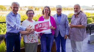 Zwei Frauen und drei Männer stehten draußen. Im Hintergrund sieht man eine Fluss. Eine Frau hält ein Schild in der Hand mit der Aufschrift "Ja zum Alter!"