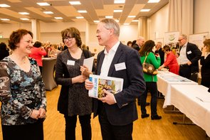 Zwei junge Frauen und ein älterer Mann stehen in einem Raum in den Stehtische zu sehen sind aber auch Stände. Sie halten Broschüren in der Hand und unterhalten sich. Klick öffnet eine vergrößerte Ansicht.