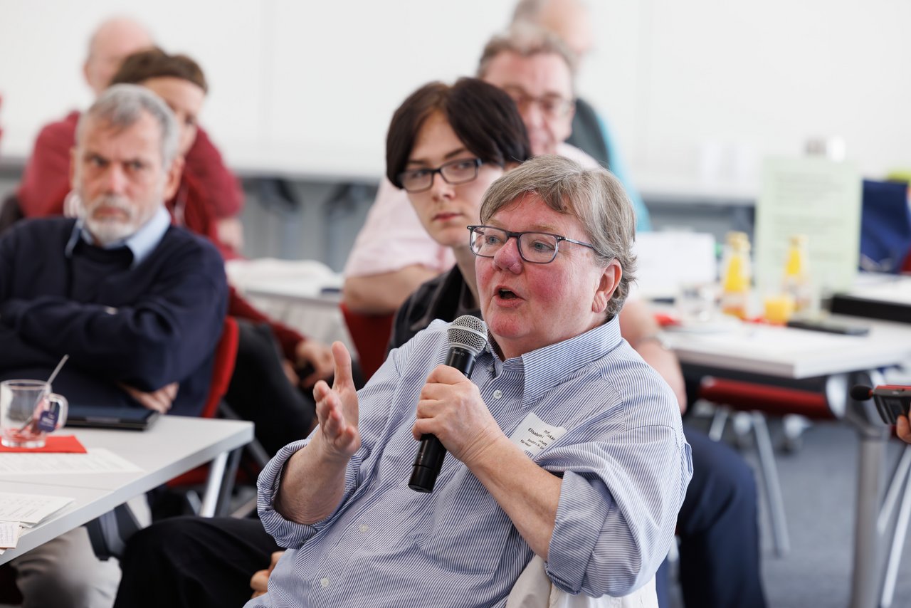 Ältere Frau mit kurzen Haaren und einer Brille spricht gestikulierend in ein Mikrofon. Im Hintergrund sind Menschen unterschiedlichen Alters an Tischen zu sehen, die ihr zuhören.