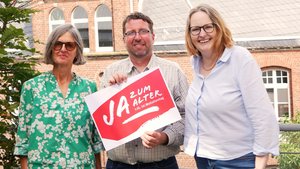 Zwei Frauen und ein Mann stehen im Freien. Der Mann hält ein Schild in der Hand mit der Aufschrift "Ja zum Alter!"