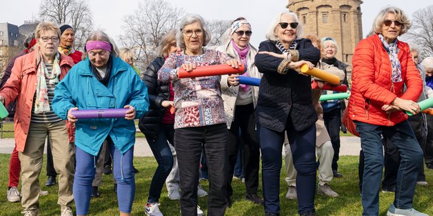 ©BAGSO_Lys Ältere Frauen stehen in einem Park. Sie haben in der Hand bunte Rollen und machen damit Gymnastik. Sie haben alle offensichtlich Spaß.