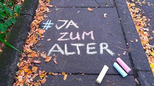 ©BAGSO/Münk Auf dem Boden steht mit bunter Kreide geschrieben: "#JA zum Alter 1.1025 Weltaltentag." Unter dem Schriftzug liegen drei bunte Kreidestücke.
