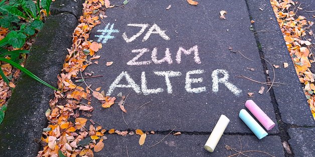 ©BAGSO/Münk Auf dem Boden steht mit bunter Kreide geschrieben: "#JA zum Alter 1.1025 Weltaltentag." Unter dem Schriftzug liegen drei bunte Kreidestücke.