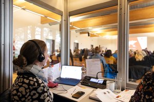 Eine Dolmetscherin mit Kopfhörern sitzt in einer Dolmetscherkabine. Vor ihr sind ein Laptop, die Verdolmetschungsanlage und verschiedene Unterlagen zu sehen. Durch die Glaswand der Kabine kann man in den Veranstaltungsraum mit dem Publikum sehen. Klick öffnet eine vergrößerte Ansicht.