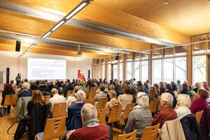 ©BAGSO_Seng Participants at BAGSO conference follow a lecture in a bright conference room. On the left in the foreground is a speaker at the lectern, on the right is a BAGSO banner. Klick öffnet eine vergrößerte Ansicht.