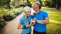 © Emir Memedovski_istock Ein älterer Mann und eine Frau stehen in einem Park. Sie haben beide Sportsachen an und tragen Kopfhörer. Die Frau hat ein Handy in der Hand und ihre Hand auf die Schulter des Mannes gelegt. Er trägt wahrscheinlich eine Smartwatch und zeigt auf das Zifferblatt.