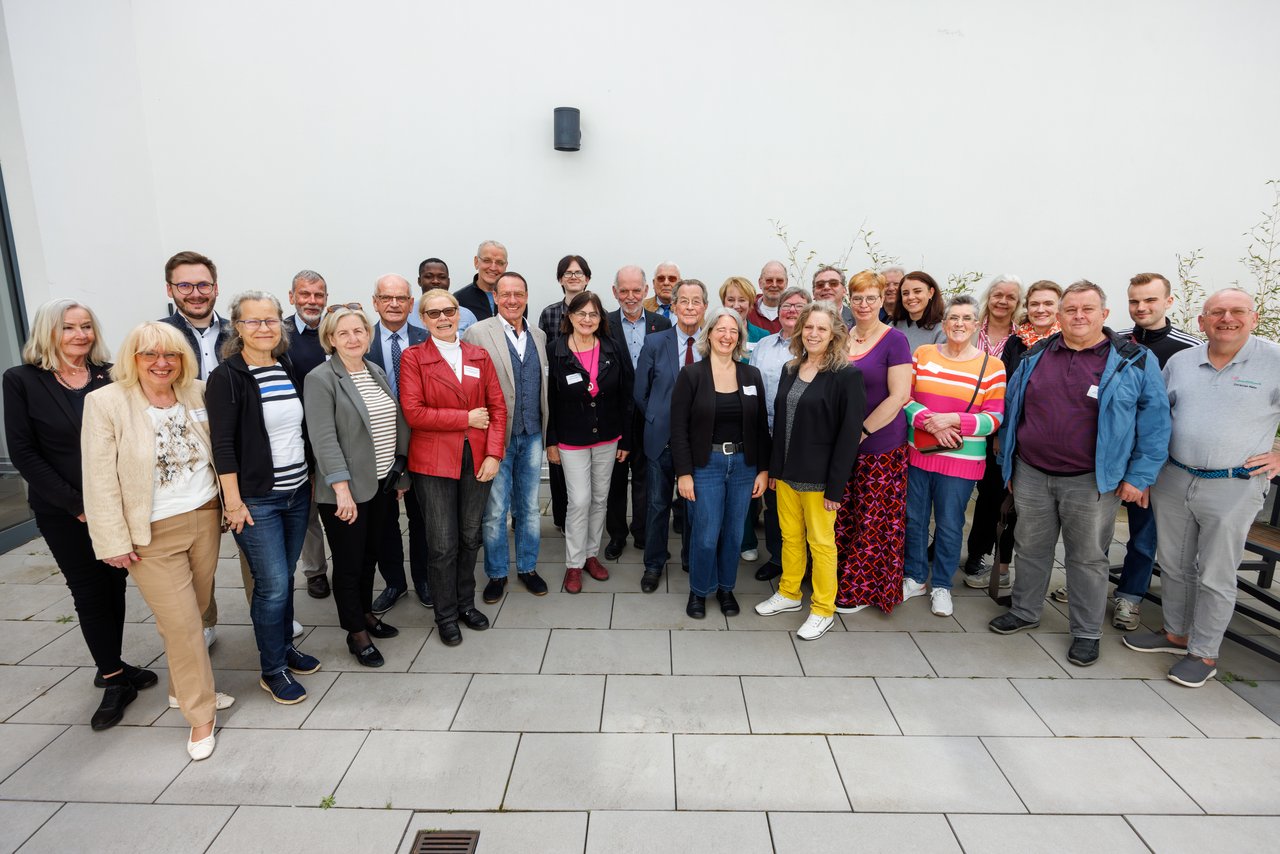 Eine große Gruppe von Menschen verschiedenen Alters stehen auf einer großen Terrasse. Fast alle schauen lächeln.