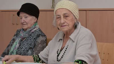 Zwei alte Damen, die beide einen Hut aufhaben, sitzen an einem Tisch. Sie haben einen Teller vor sich.