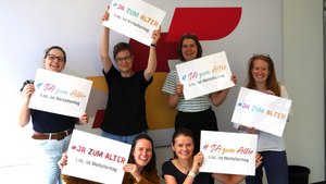 ©BAGSO Sechs Frauen stehen vor einer Fotowand, die BAGSO-Logo-Elemente zeigt. Sie lachen alle und halten jeweils ein Schild in der Hand mit der Aufschrift "Ja zum Alter!"