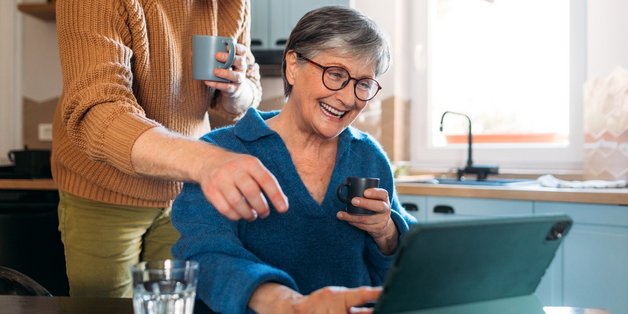 Eine ältere Frau mit kurzem grauen Haar, Brille und blauem Pullover sitzt am Küchentisch. Über ihr steht ein junger Mann mit Rollkragenpullower. Sie haben beide eine Tasse in der Hand und schauen auf ein Tablet, dass auf dem Tisch steht. Beide zeigen auf den Bildschirm. 