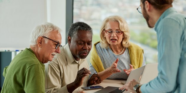 ©mmillan_istock Eine ältere Frau und 3 ältere Männer sitzen um einen Tisch. Darauf steht ein Laptop und ein Smarphone. Sie schauen auf den Bildschirm. Vor ihnen steht eien Mann, der ihnen in einem Buch etwas zeigt.