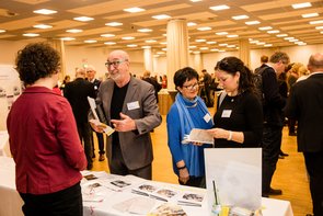 Blick einen großen Raum, in dem Messestände zu sehen sind. An einem Stand sieht man einen älteren Mann, der sich mit einer jungen Frau, die hinter dem Stand steht, unterhält. Neben ihm stehen zwei Frauen, die sich miteinander unterhalten. Klick öffnet eine vergrößerte Ansicht.