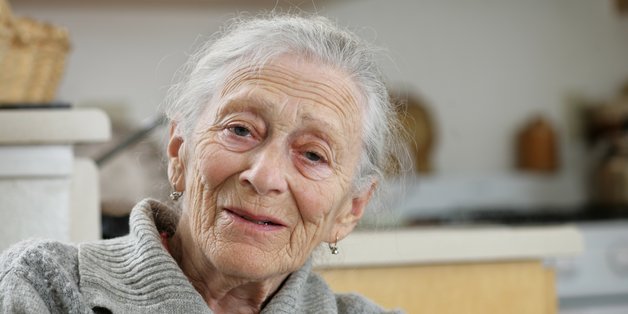ogoboom/stock.adobe.com Ältere Frau mit weißem Haar und grauem Strickpullover sitzt in einer gemütlichen Küche und lächelt freundlich in die Kamera.