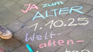 © BAGSO/Münk Man sieht links im Bild zwei Füße, die Turnschuge tragen. Davor liegen bunte Kreidestücke. Auf dem Boden steht "JA zum Alter 1.1025 Weltaltentag."