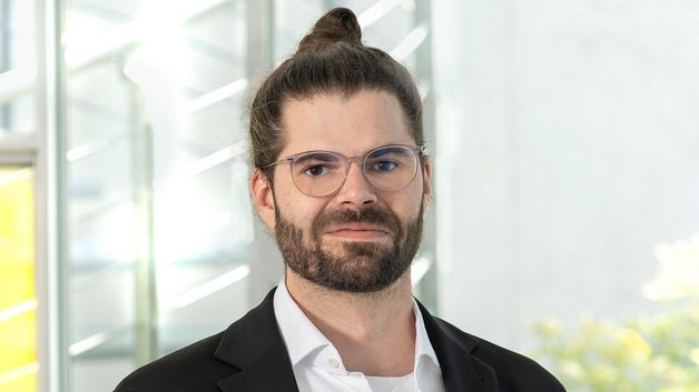 ©BAGSO/Sachs Portrait Alexander Engel: Junger Mann, mit Brille und Bart. Er hat seine schwarzen langen Haare zu einem Knoten hochgebunden. Er trägt ein weißes Hemd und ein schwarzes Sakko.