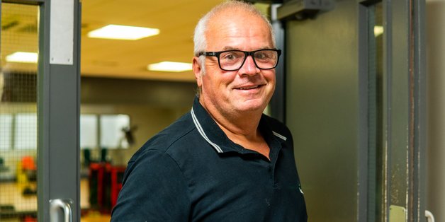 Ein älterer Mann macht gerade eine Tür auf. Im Hintergrund sieht man eine Werkshalle.