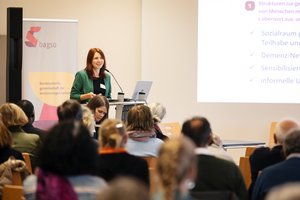 Die Referentin Ute Hauser, eine junge Frau mit langem, rotem Haar, steht an einem Rednerpult. Sie trägt einen grünen Blazer. Hinter ihr ist ein Banner der BAGSO zu sehen. Im Vordergrund sitzen Teilnehmende an der Fachtagung. Klick öffnet eine vergrößerte Ansicht.