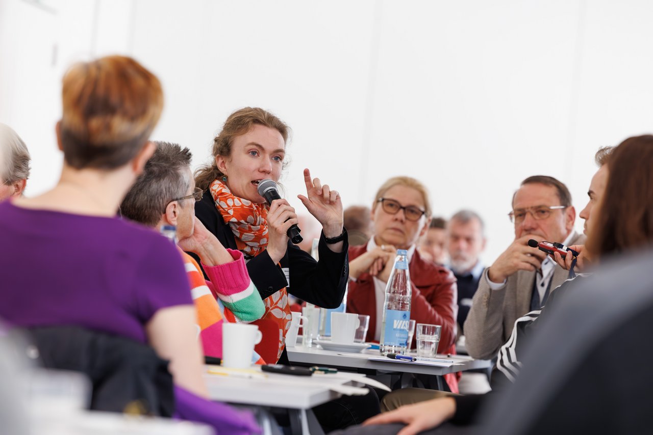 Jüngere Frau spricht in ein Mikrofon. Sie sitzt an einem Tisch, um sie rum sind andere Tagungs-Teilnehmende zu sehen, die ihr zuhören.