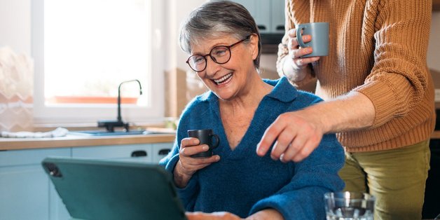 Eine ältere Frau mit kurzem grauen Haar, Brille und blauem Pullover sitzt am Küchentisch. Über ihr steht ein junger Mann mit Rollkragenpullower. Sie haben beide eine Tasse in der Hand und schauen auf ein Tablet, dass auf dem Tisch steht. Beide zeigen auf den Bildschirm. 